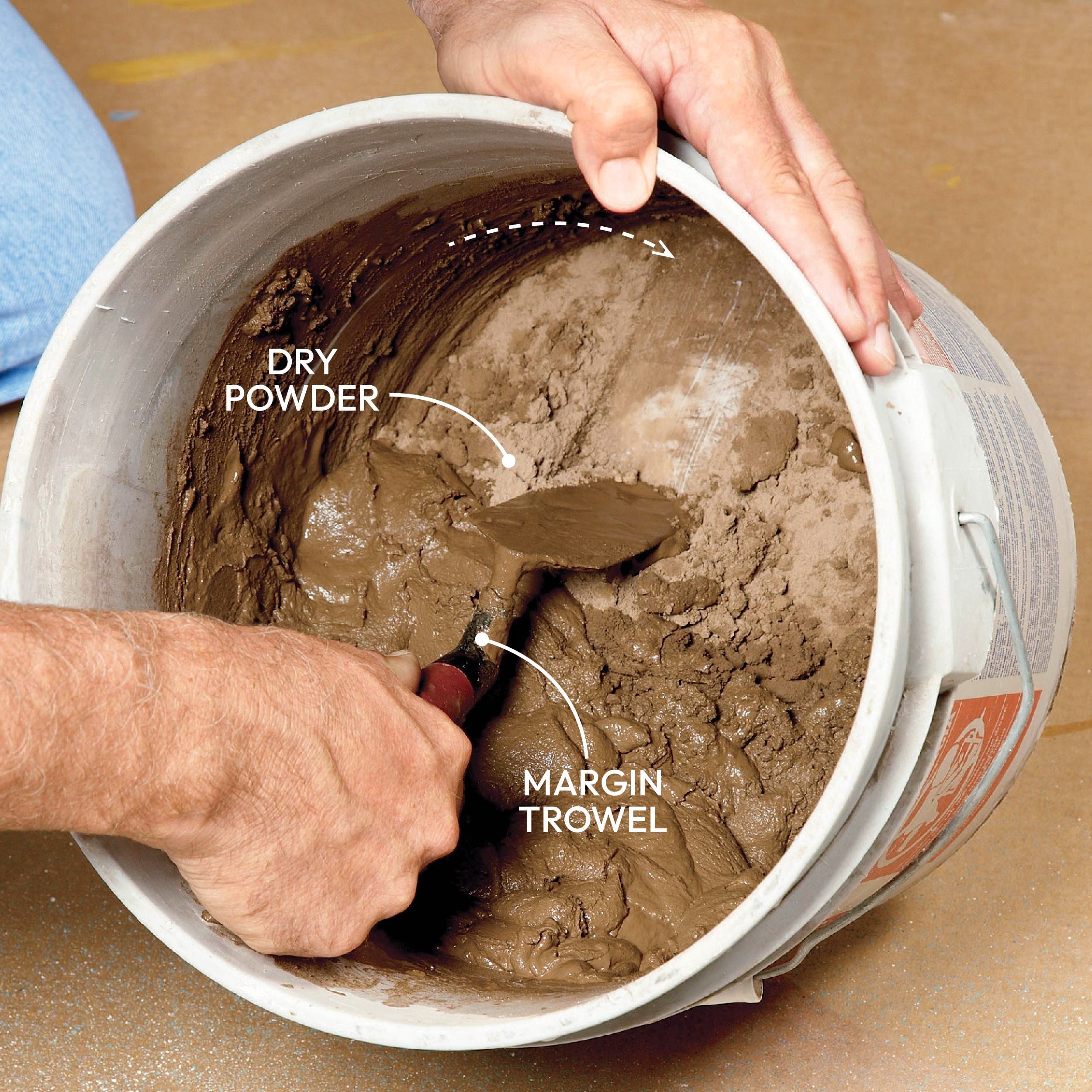 A hand holds a margin trowel, mixing wet clay in a bucket containing dry powder, on a floor with visible texture.