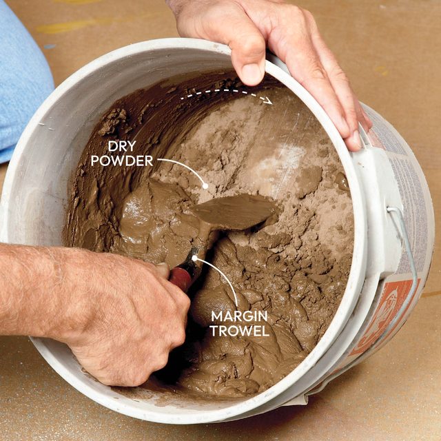 A hand holds a margin trowel, mixing wet clay in a bucket containing dry powder, on a floor with visible texture.
