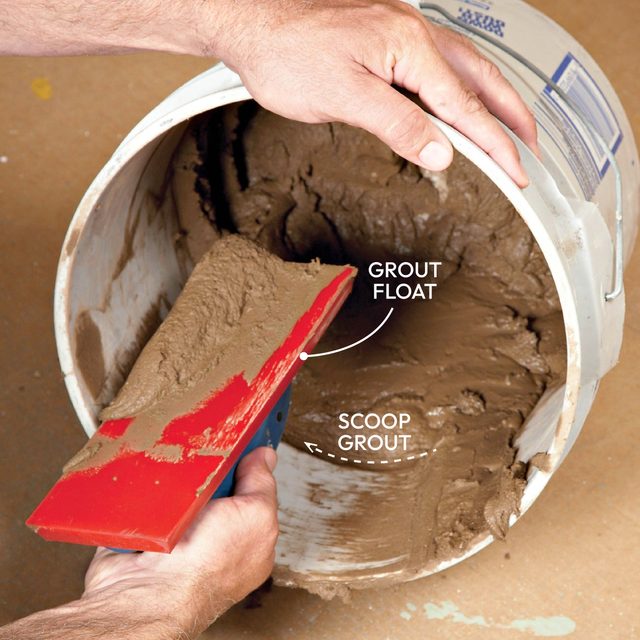 A hand holds a red grout float above a bucket filled with wet grout, preparing to scoop some out for use in tiling.