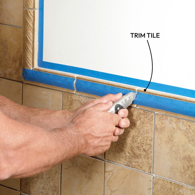 A hand uses a utility knife to trim tile along a blue painter's tape strip, with textured floor tiles visible below.