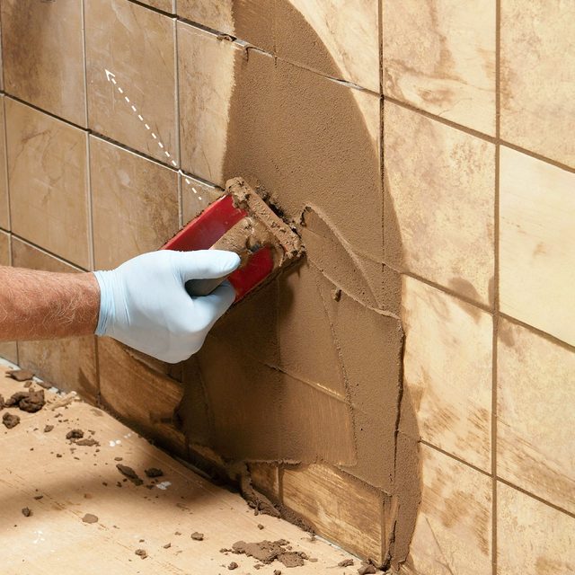 A hand wearing a glove smooths tile adhesive with a tool against a tiled wall, amidst a construction or renovation setting.