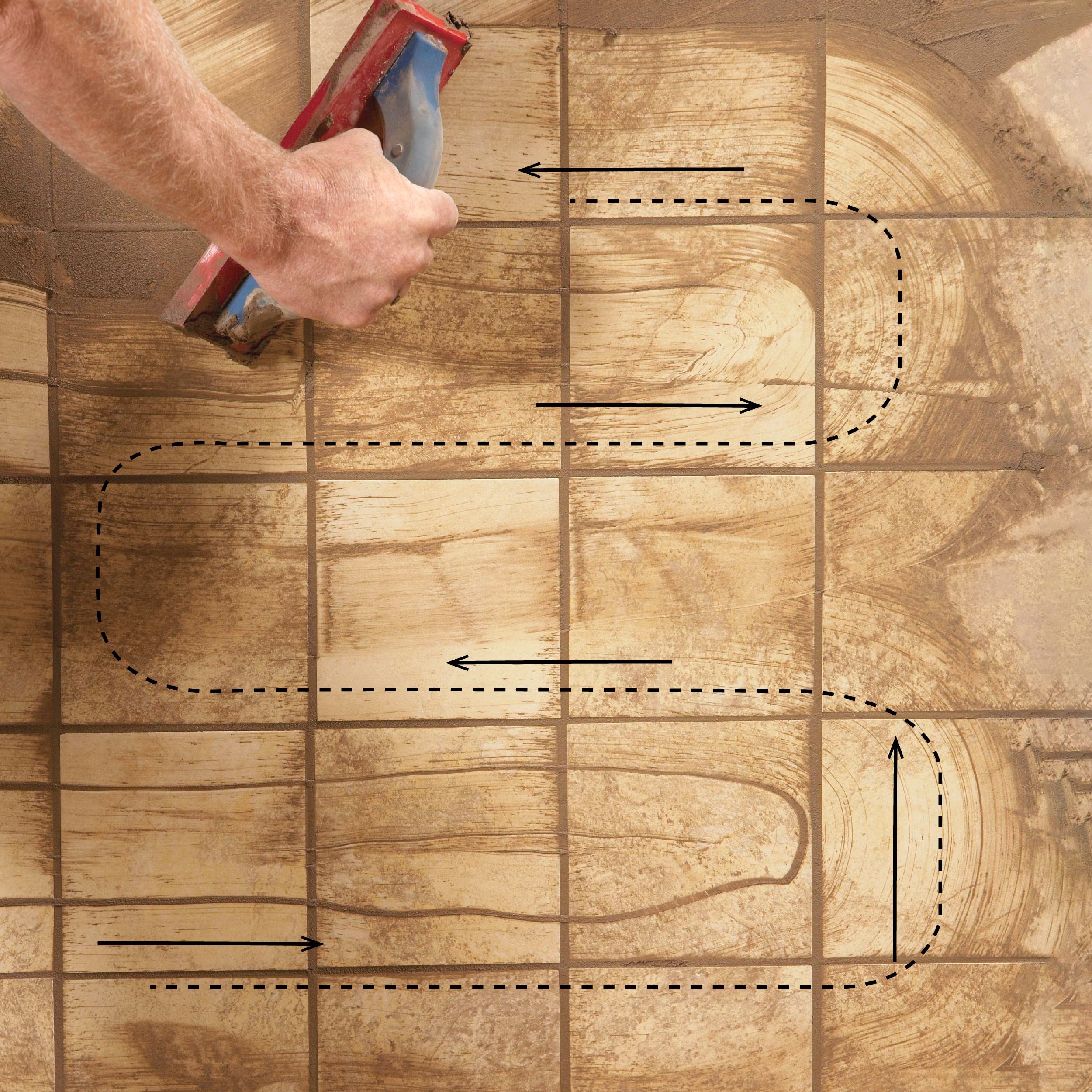 A hand holds a smoothing tool, applying a brown substance in curved, patterned strokes over a textured surface, resembling tiled flooring.