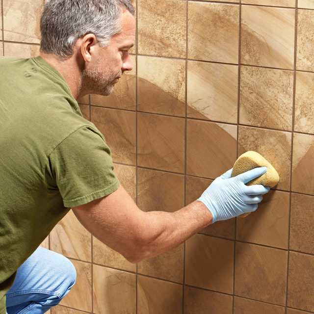 A man wearing blue gloves scrubs a tiled wall with a yellow sponge, cleaning it in a brightly lit interior space.