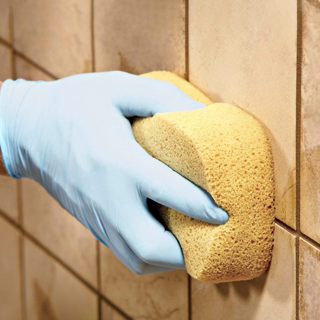 A gloved hand scrubs a sponge against a tiled wall, removing dirt or stains in a bathroom or kitchen environment.