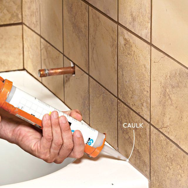 A hand is applying caulk between tiled surfaces near a bathtub, ensuring a waterproof seal along the joints.