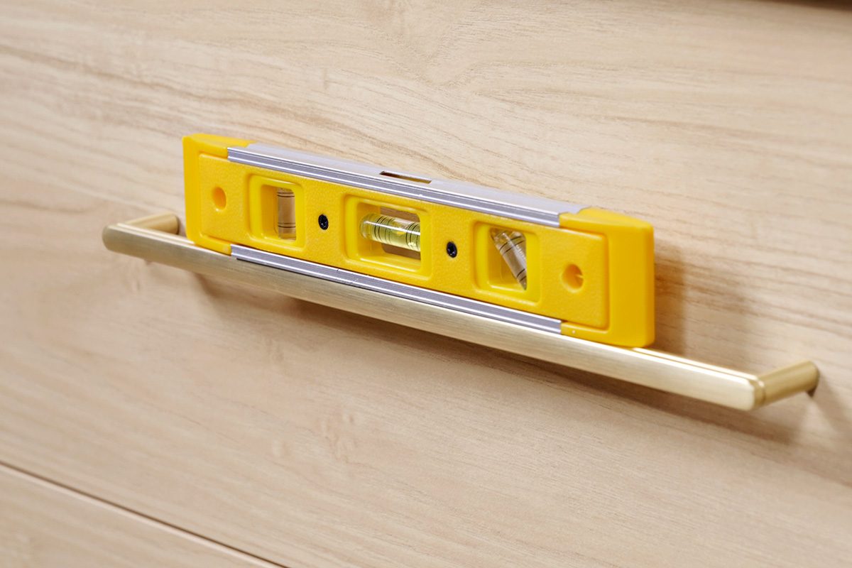 A yellow spirit level placed on a silver drawer handle attached to a light wood cabinet, showing the handle is properly aligned and level.