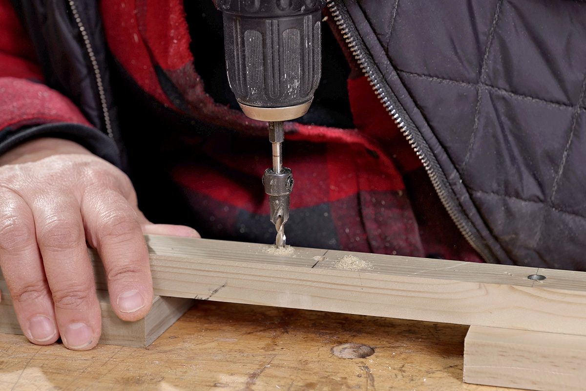 A person uses a power drill to make a hole in a piece of light-colored wood, holding the wood steady on a workbench with their hand.