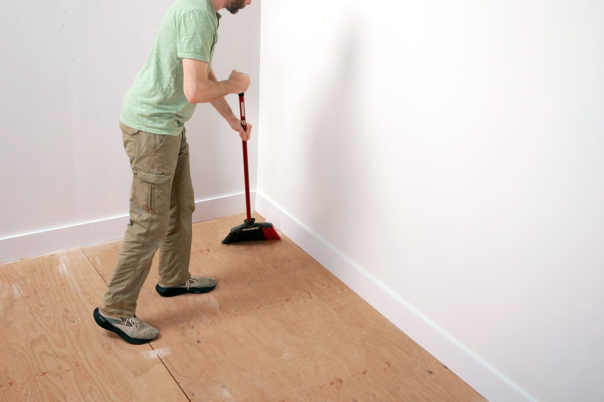 A person wearing a green shirt and khaki pants sweeps dust and debris from a bare plywood floor in a mostly empty, white-walled room.
