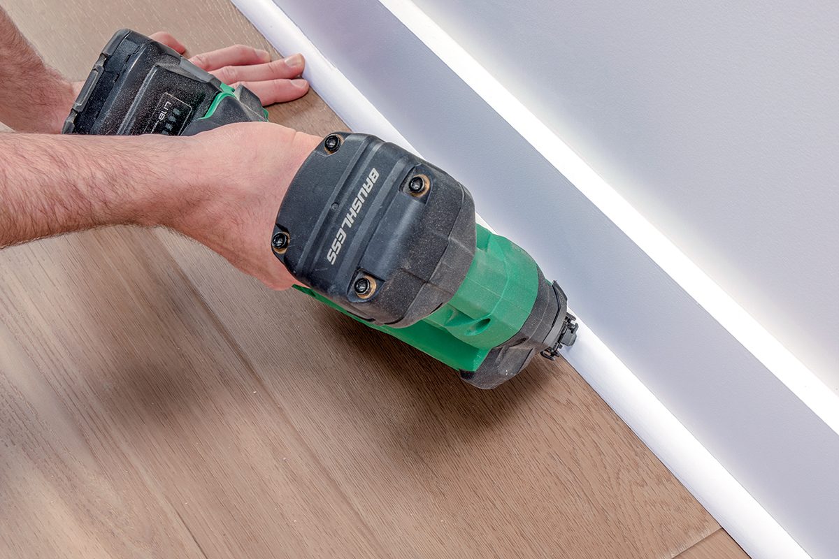 A person uses a green cordless nail gun to attach white baseboard trim to a wall above a wooden floor.