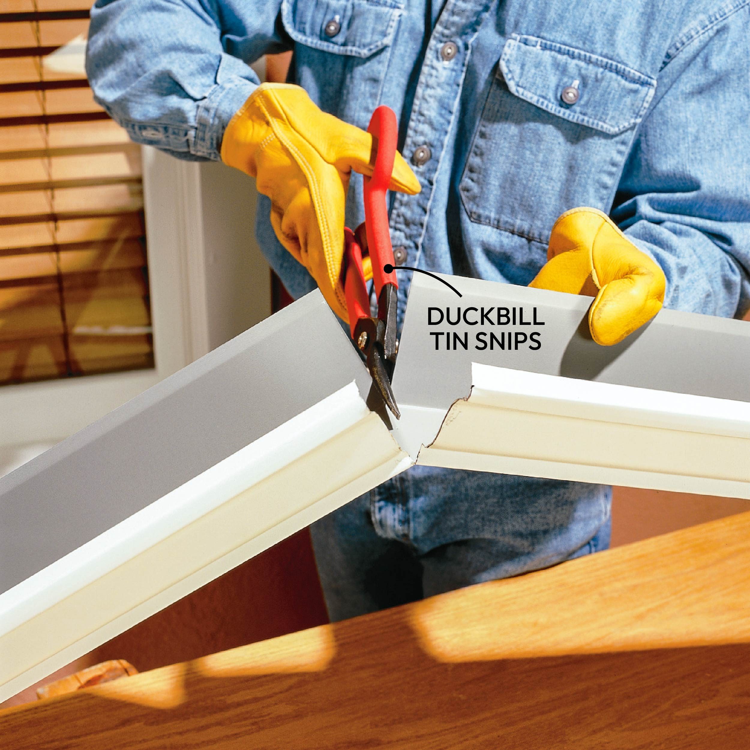 A person wearing yellow gloves uses red tin snips to cut a metal piece in a home environment.