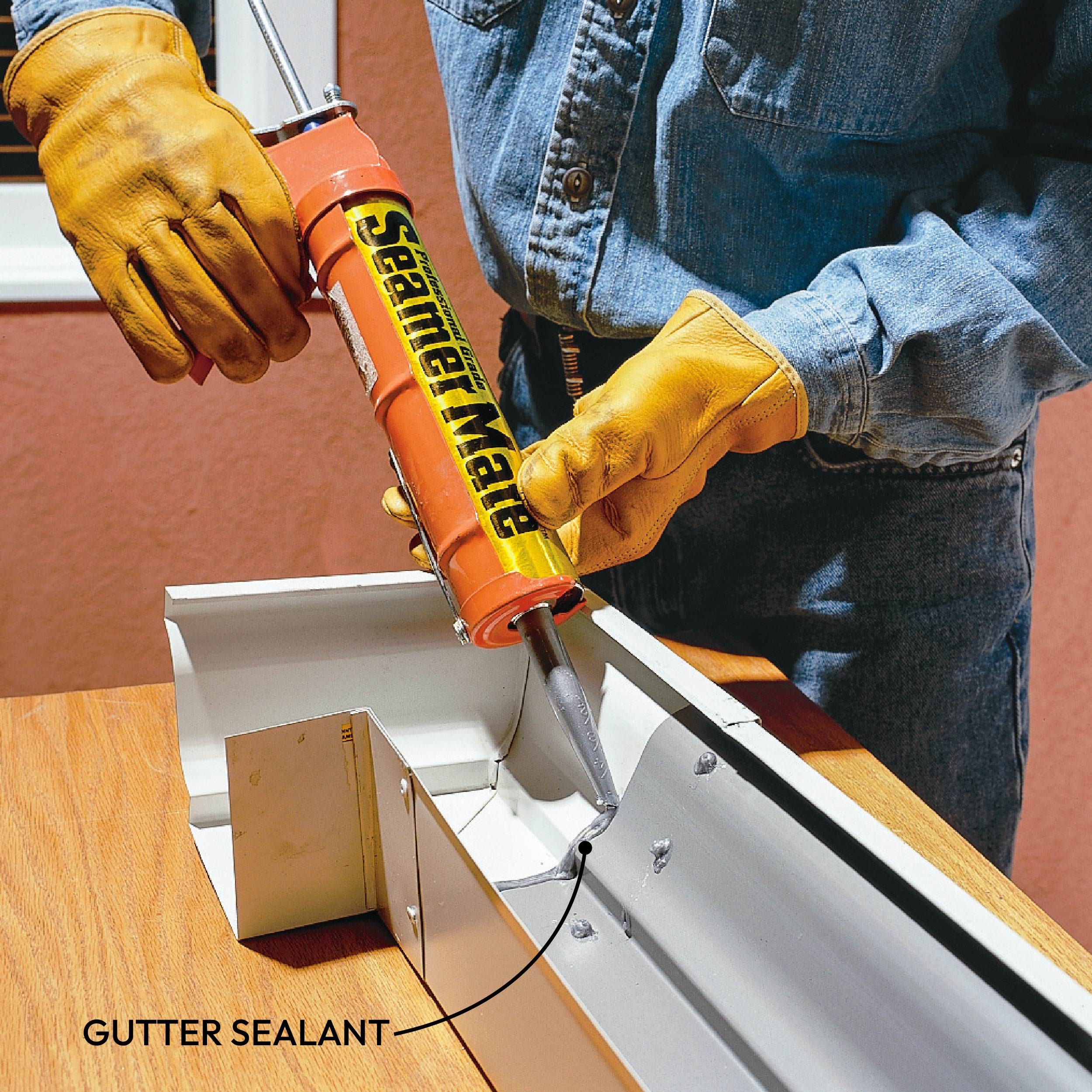 A person in gloves uses a caulking gun to apply gutter sealant into a metal gutter section on a wooden surface.