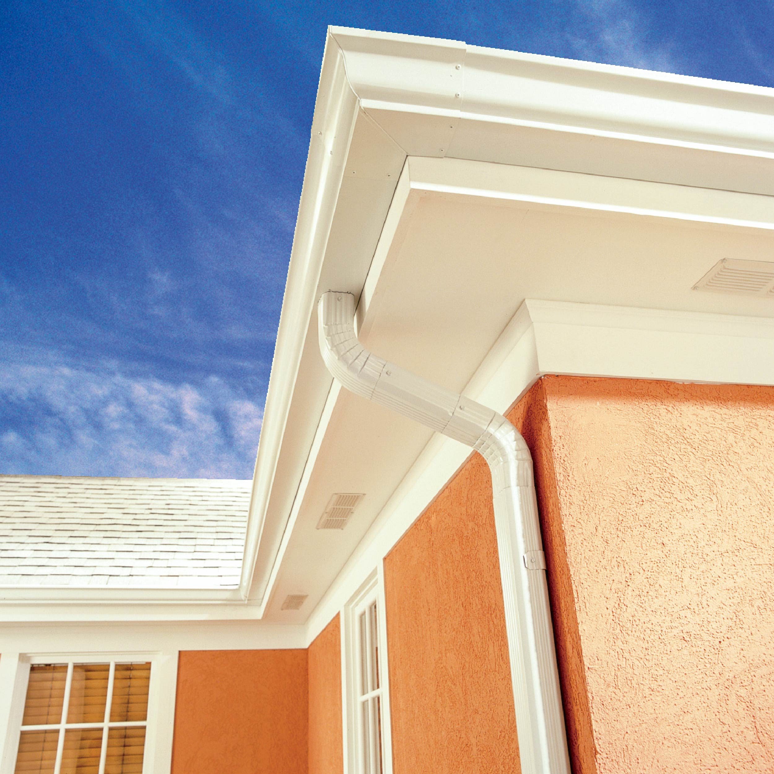 A white gutter channels water along the roof's edge of an orange building, beneath a clear blue sky. Light reflects off the surfaces.