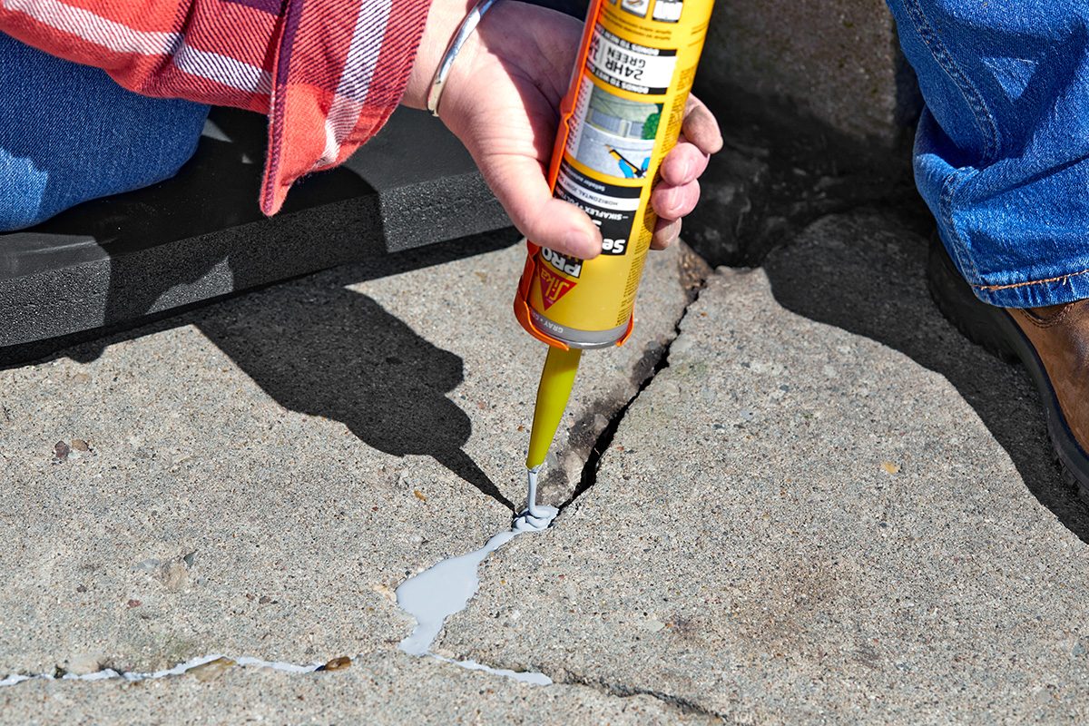 A person in a red plaid shirt applies sealant with a caulking gun to fill a large crack in a concrete surface.