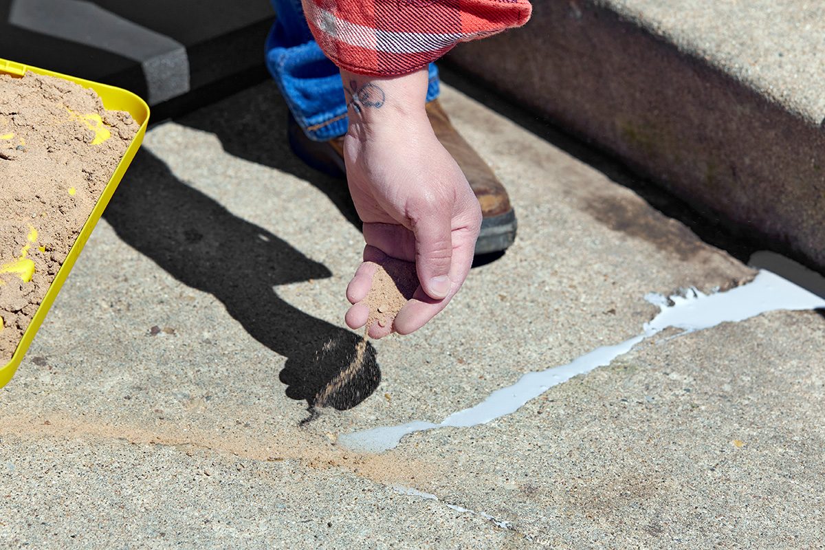 A person sprinkles sand from their hand onto wet crack filler on a concrete surface, with a tray of sand nearby and the person's arm partially visible.