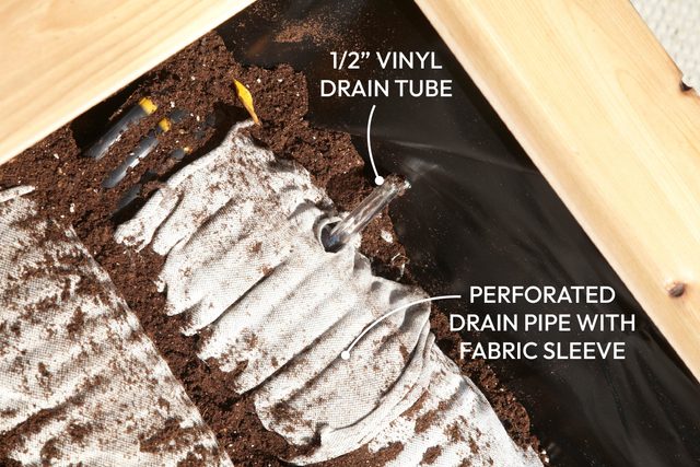 A drain pipe with a fabric sleeve is positioned in soil, indicating an irrigation setup for gardening in a wooden planter.