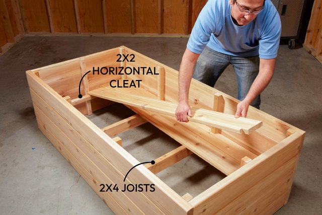 A person is positioning a wooden cleat inside a partially constructed wooden box in a garage-like space with wooden walls and a concrete floor.