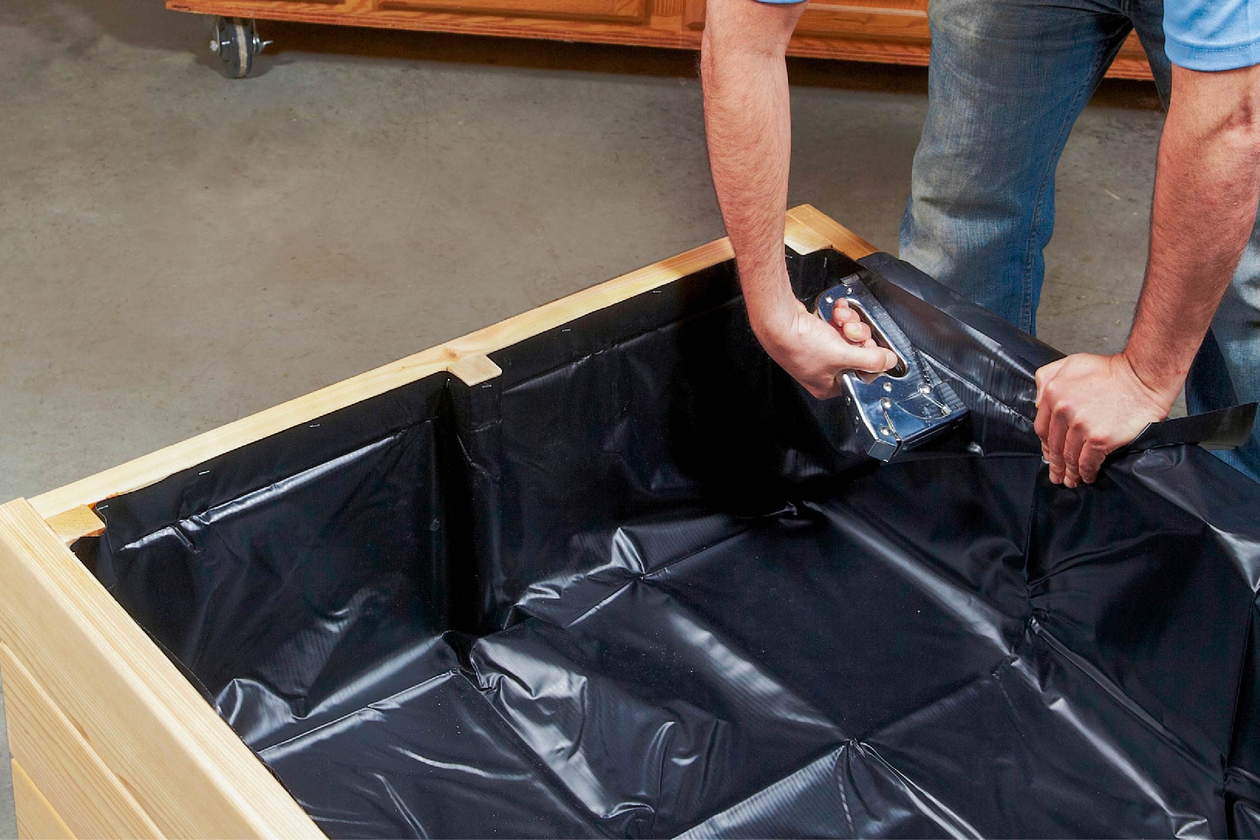A person attaches a black plastic liner inside a wooden container, working in a garage setting with cabinetry in the background.