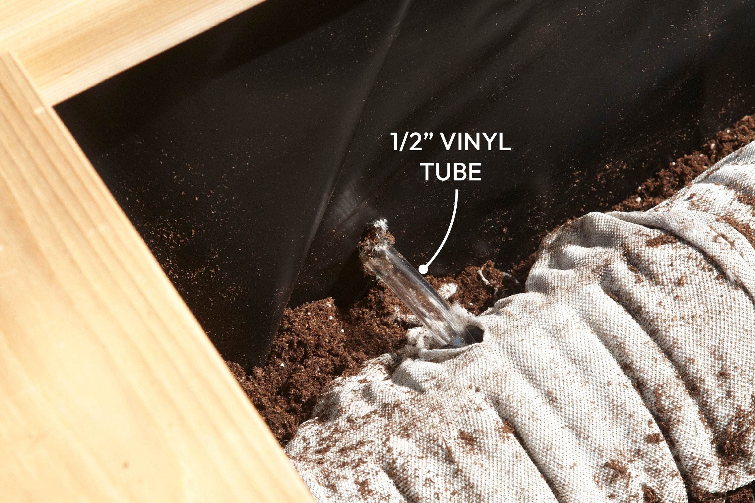 A vinyl tube releases water into soil within a raised garden bed, surrounded by black plastic and wooden edging, enhancing irrigation for plant growth.