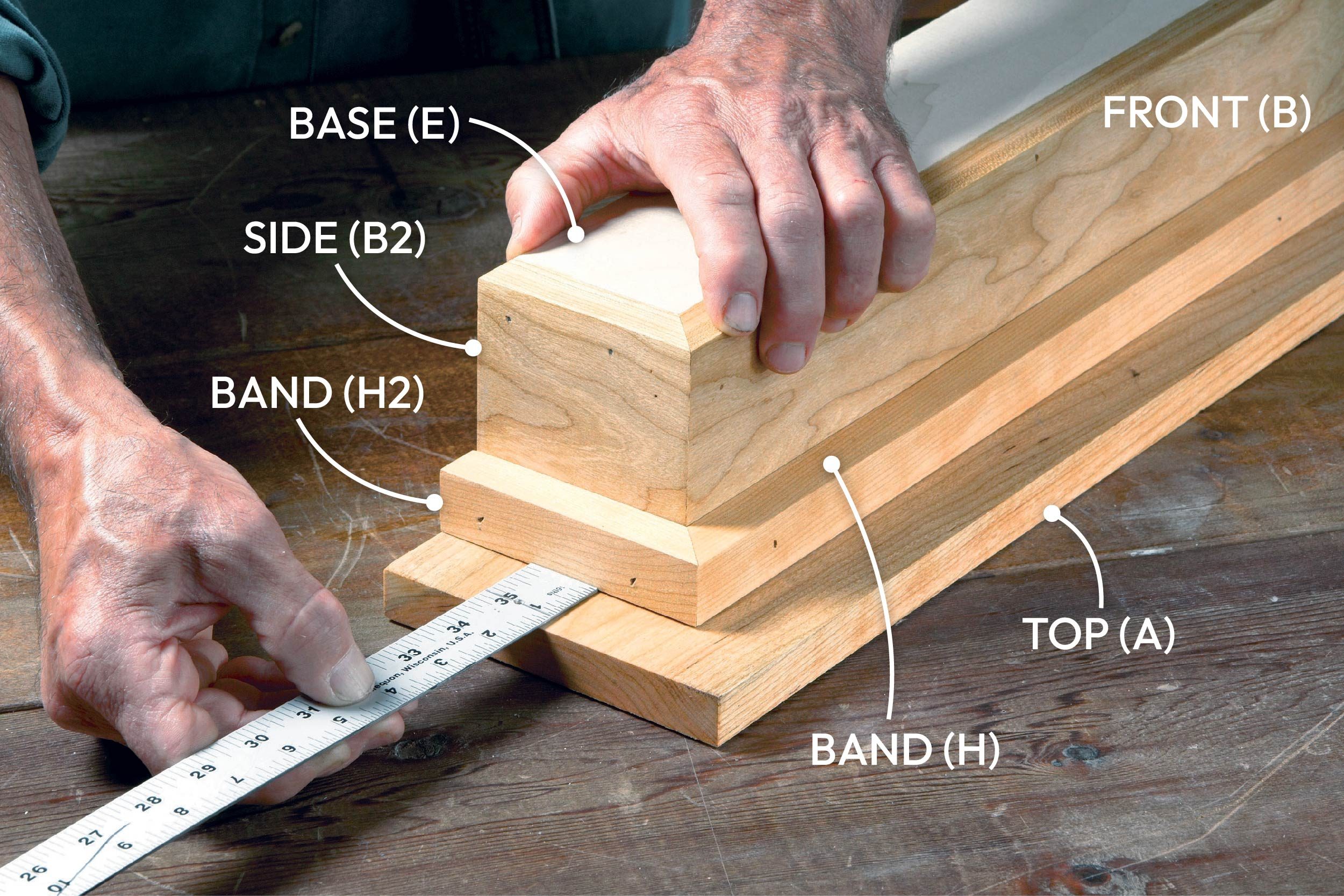 A hand holds a measuring tape against wooden pieces labeled with their respective parts, while another hand supports a stacked wooden structure on a workbench.