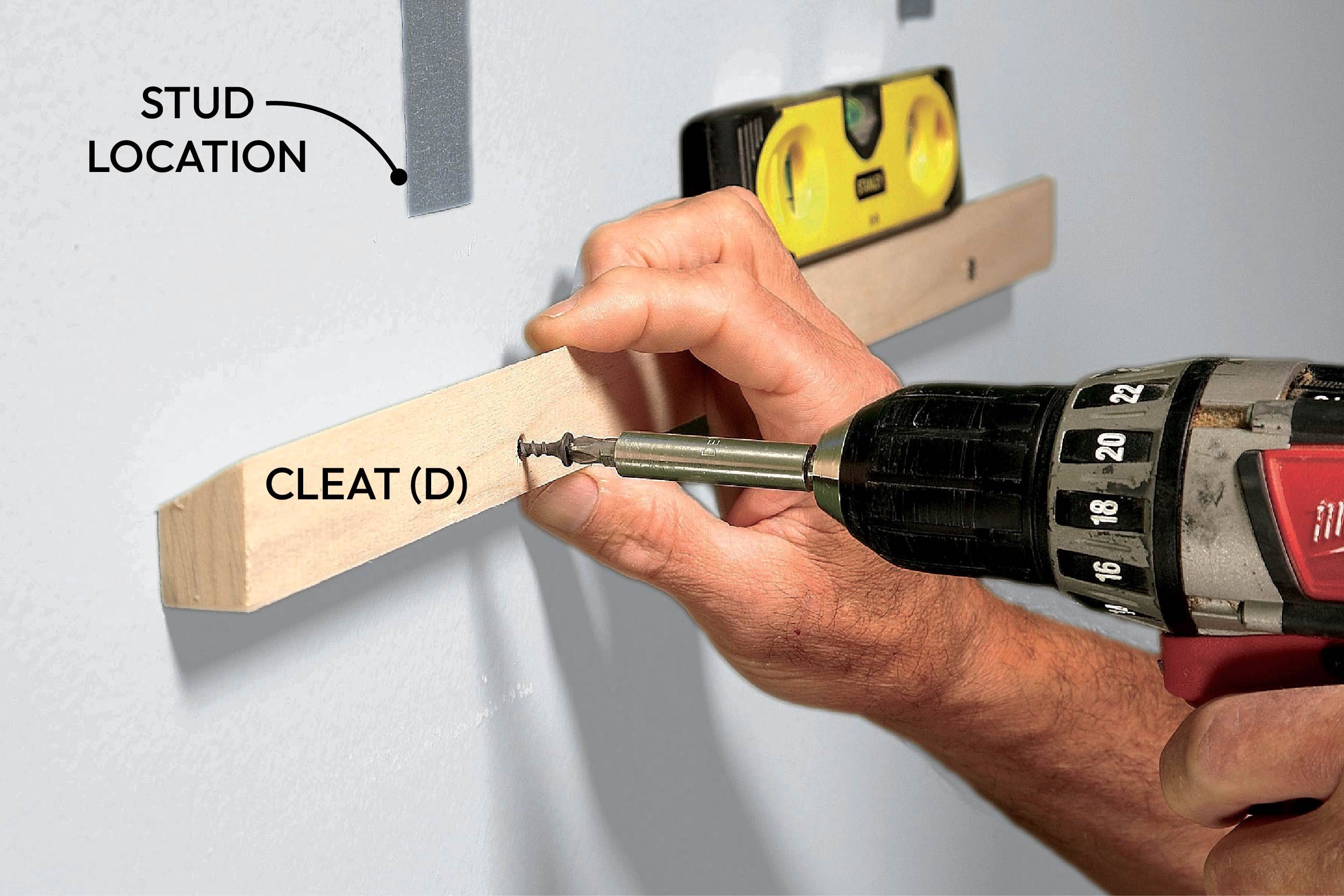 A hand drills a screw into a wooden cleat mounted on a green wall, with markings indicating stud locations and a level nearby.