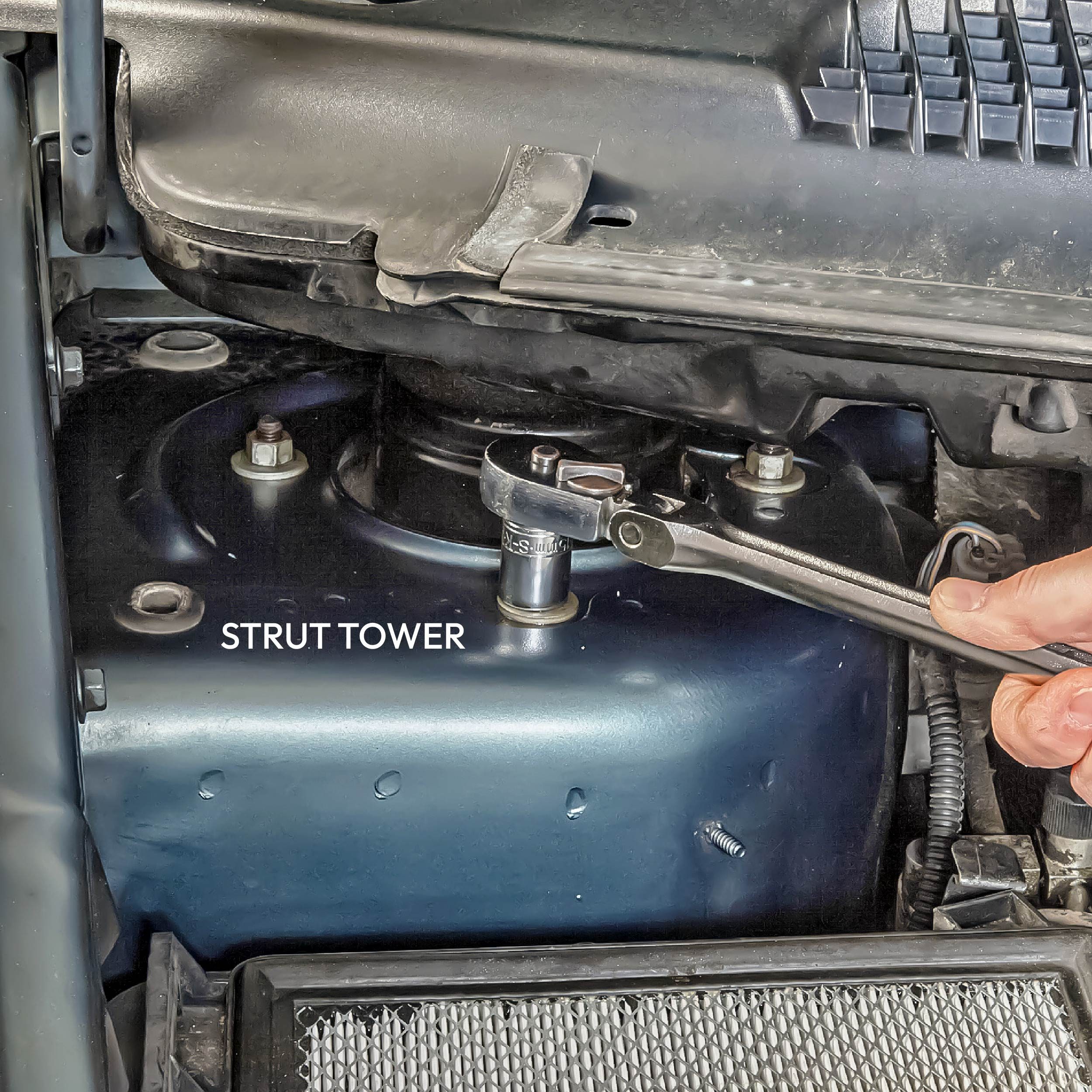 A ratchet wrench fastens a bolt on a strut tower within a vehicle&rsquo;s engine compartment, surrounded by various mechanical components and fittings.