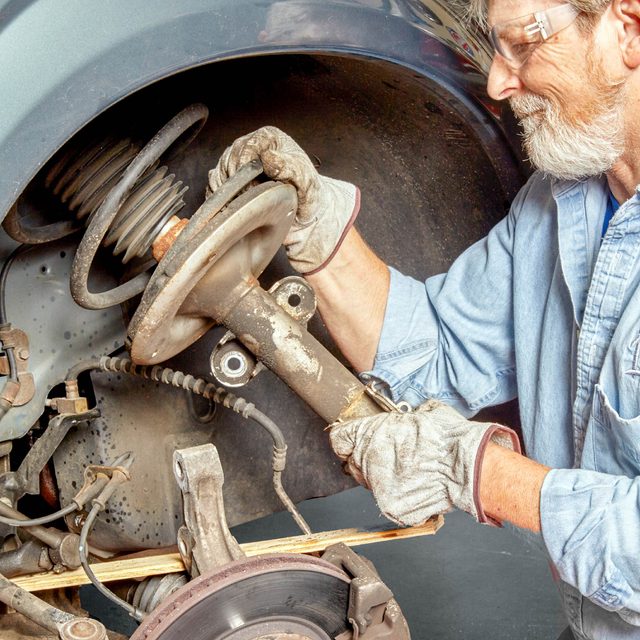 A man in gloves works on a car's suspension, removing a part near the brake assembly inside a garage.
