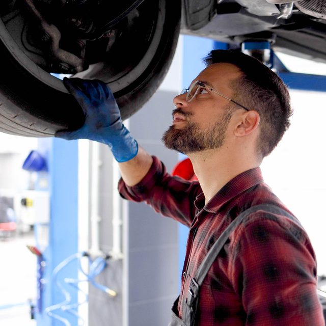 A mechanic inspects a vehicle's undercarriage, in a well-lit garage with tools and equipment visible in the background.