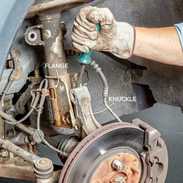 A hand wearing a glove uses a screwdriver to adjust components labeled "Flange" and "Knuckle" on a vehicle's suspension system against a dark background.