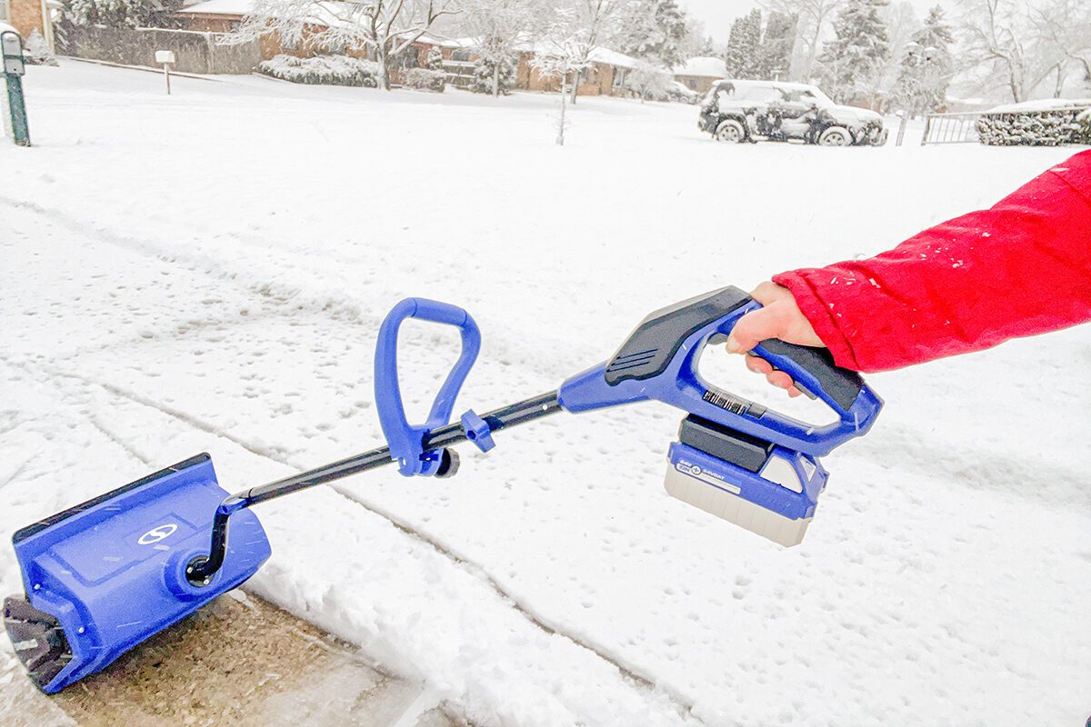 A person in a red jacket uses a blue cordless snow shovel to clear snow from a sidewalk on a snowy day; houses and a car are visible in the snowy background.