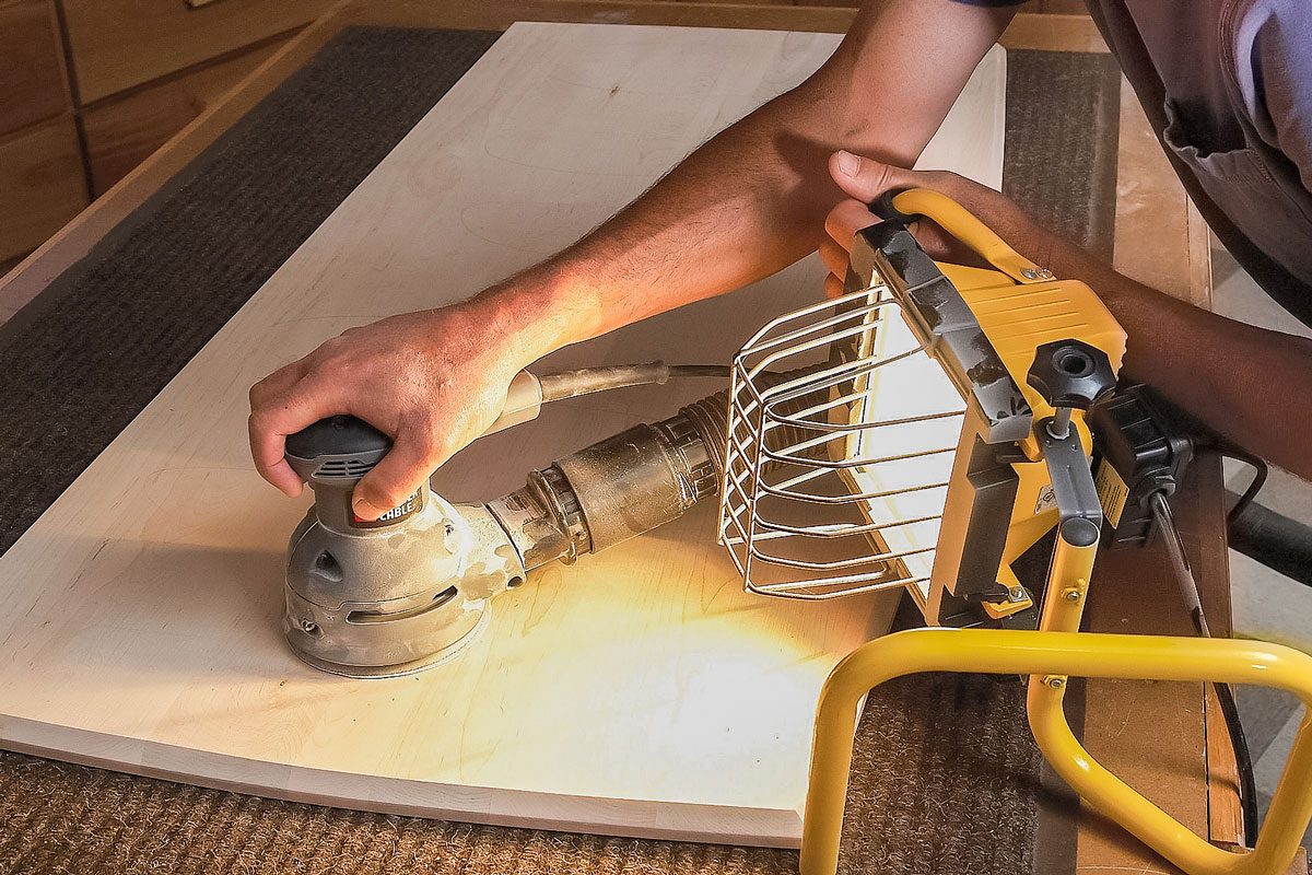 A person wearing safety glasses and red ear protection uses a power sander on a wooden board, illuminated by a bright work light on a workbench in a woodshop;