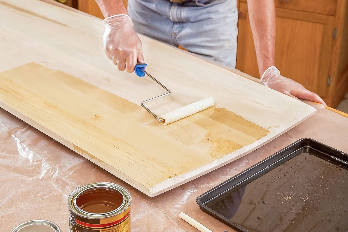 A man in a purple shirt uses a paint roller to apply wood stain to a large wooden board; The table is covered with plastic, and there are cans of stain and painting tools nearby;