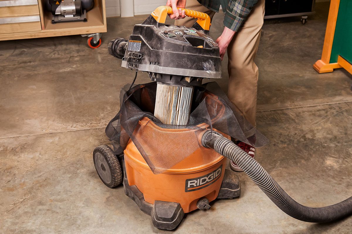 A person empties debris from a RIDGID wet/dry shop vacuum by lifting the top and using a filter bag; The vacuum is on a concrete floor in a workshop setting;