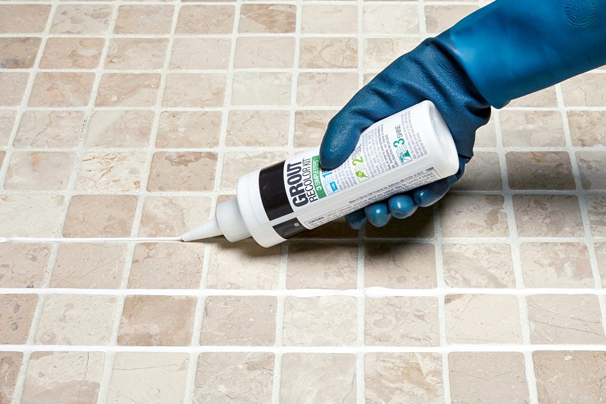 A gloved hand applies grout from a squeeze bottle to the joints between beige square tiles on a tiled surface.