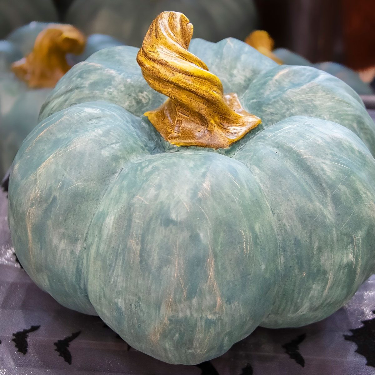 Decorative Halloween or Thanksgiving teal pumpkin with twisted stem sitting on a table with shiny bat tablecloth and other similar pumpkins blurred in background