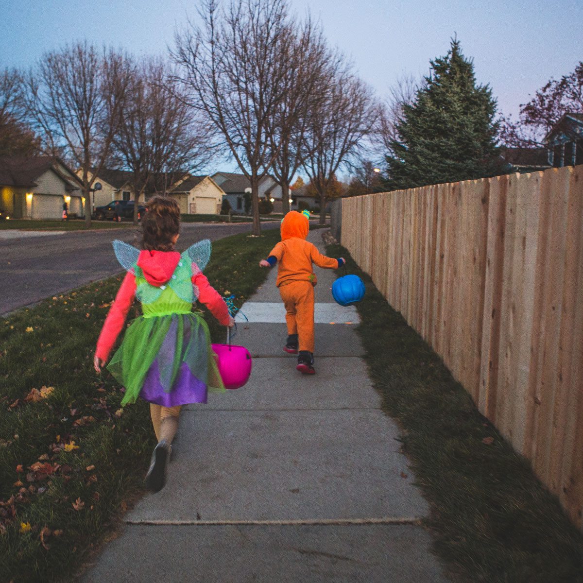 Little girl and a little boy run down a sidewalk on halloween in their halloween costumes carrying their treat buckets.