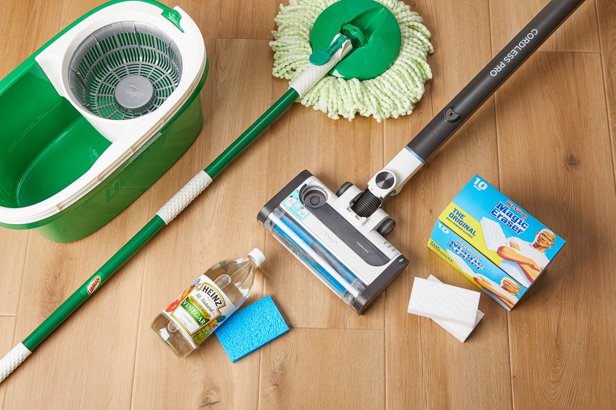 Overhead shot of household cleaning supplies arranged on a wooden floor, featuring a green mop and bucket, a cordless vacuum, a bottle of white vinegar, a blue sponge, and a box of Mr Clean Magic Eraser pads