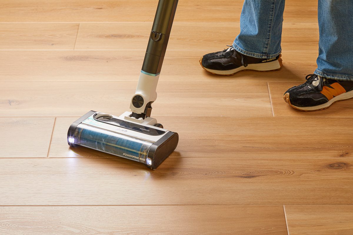 Closeup shot of a person vacuuming a wooden floor with a cordless vacuum cleaner, showing the lower legs and shoes of the person in jeans