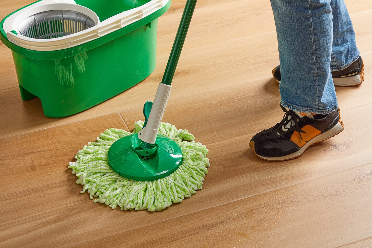 Closeup shot of a person wearing jeans and sneakers cleaning a wooden floor with a green spin mop, with a matching green mop bucket nearby