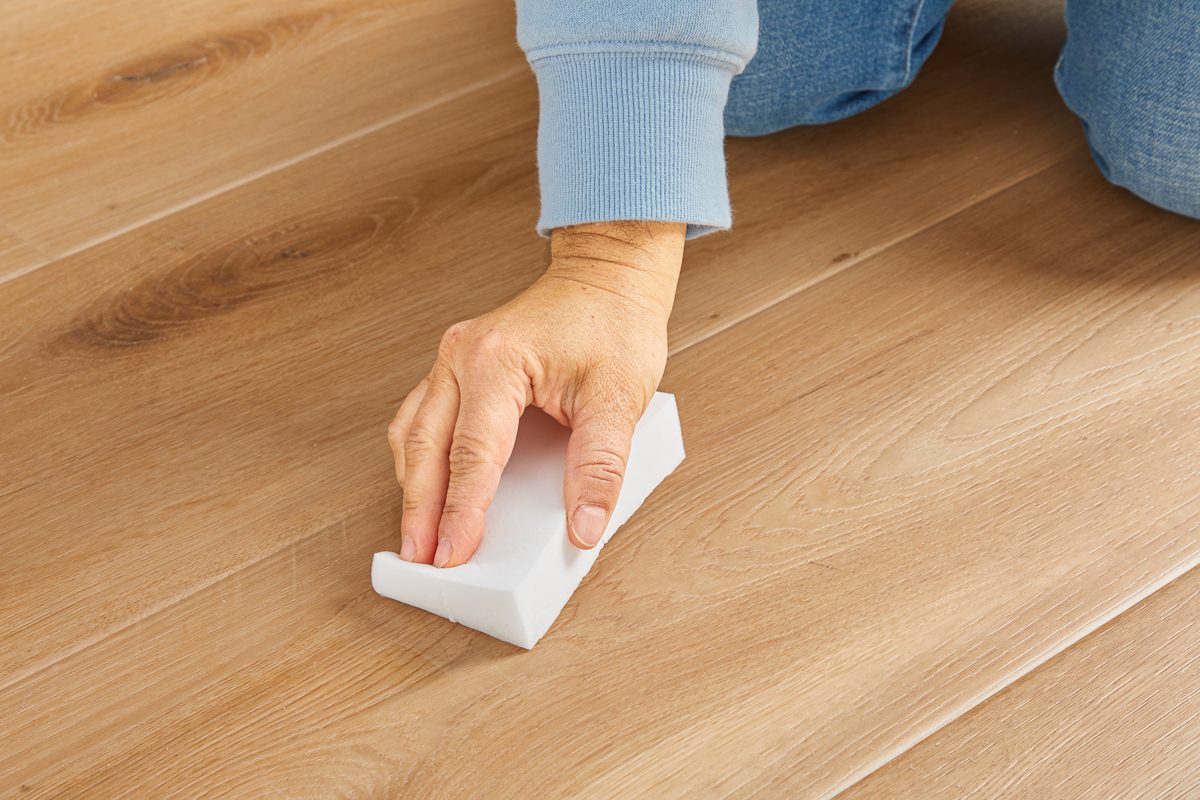 Closeup shot of a person wearing blue sleeves wiping a light wooden floor with a white cleaning sponge