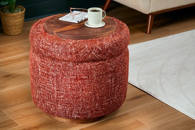 A round, red textured ottoman with a wooden tray on top, holding a cup of coffee and a book, is placed on a wooden floor next to a beige rug and a potted plant.