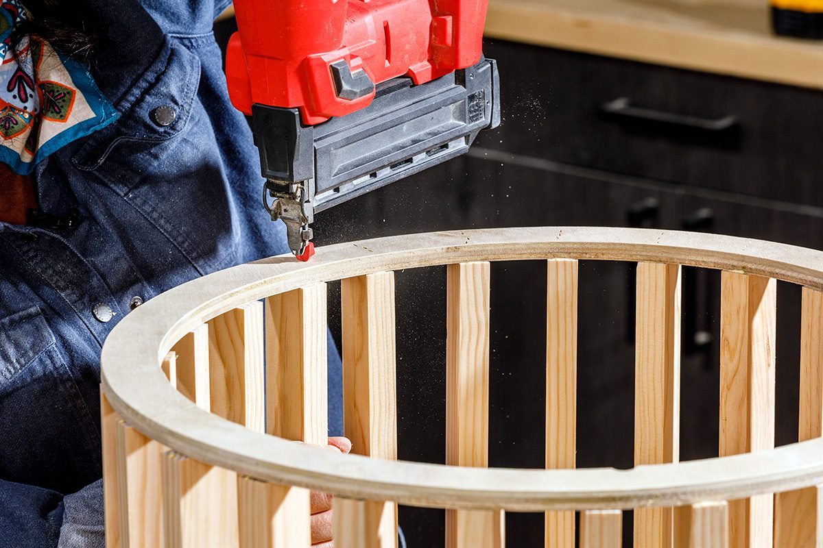 A person uses a red nail gun to fasten pieces of light wood while assembling a round wooden structure, possibly a table or a basket, in a workshop.