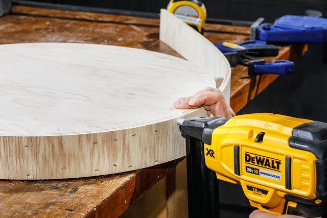 A person uses a yellow Dewalt nail gun to attach a curved wooden piece to the edge of a large, round wooden board on a workbench. Clamps and tools are visible in the background.