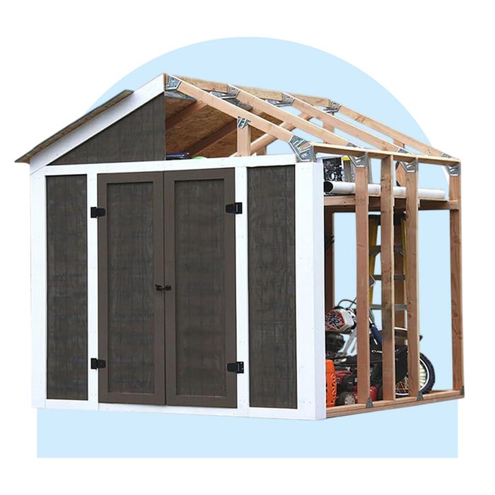 A partially constructed shed, showing one finished side with double doors and the other side exposed, revealing wooden framing and tools inside.