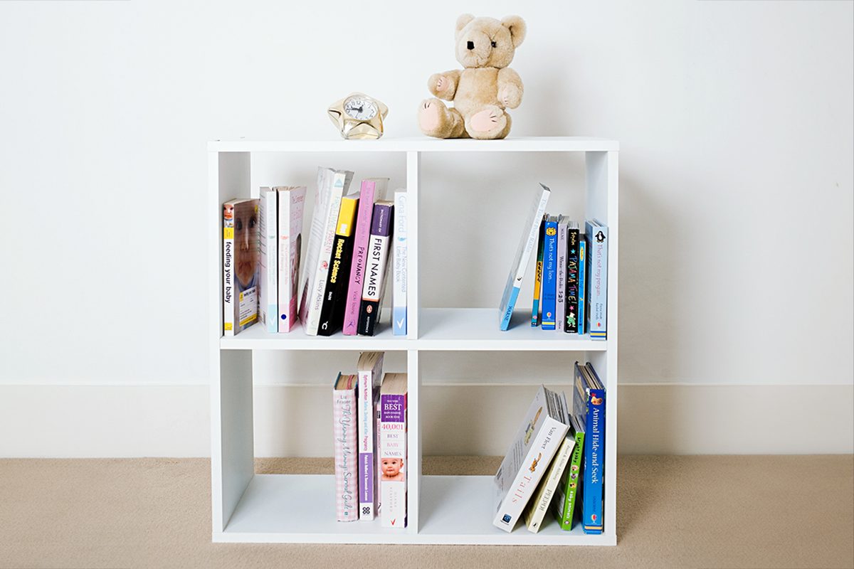A white, four-cube bookshelf holding assorted books, with a small teddy bear and a decorative ceramic animal placed on top. The background is a plain white wall and beige carpet.