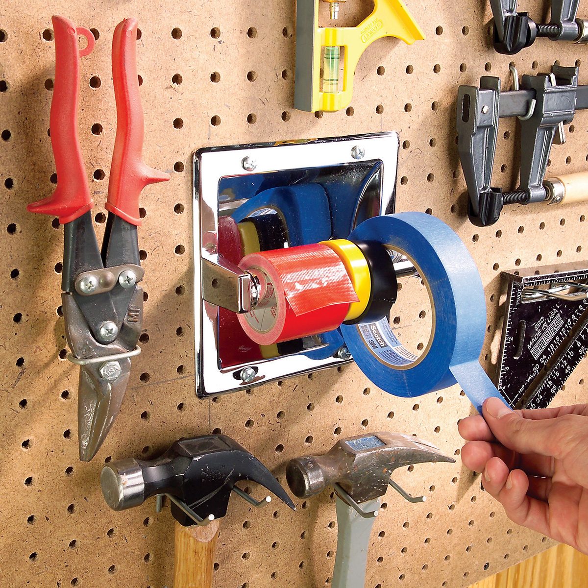 A hand pulls blue painter’s tape from a mounted tape dispenser on a pegboard wall, surrounded by tools including pliers, hammers, clamps, and a square.