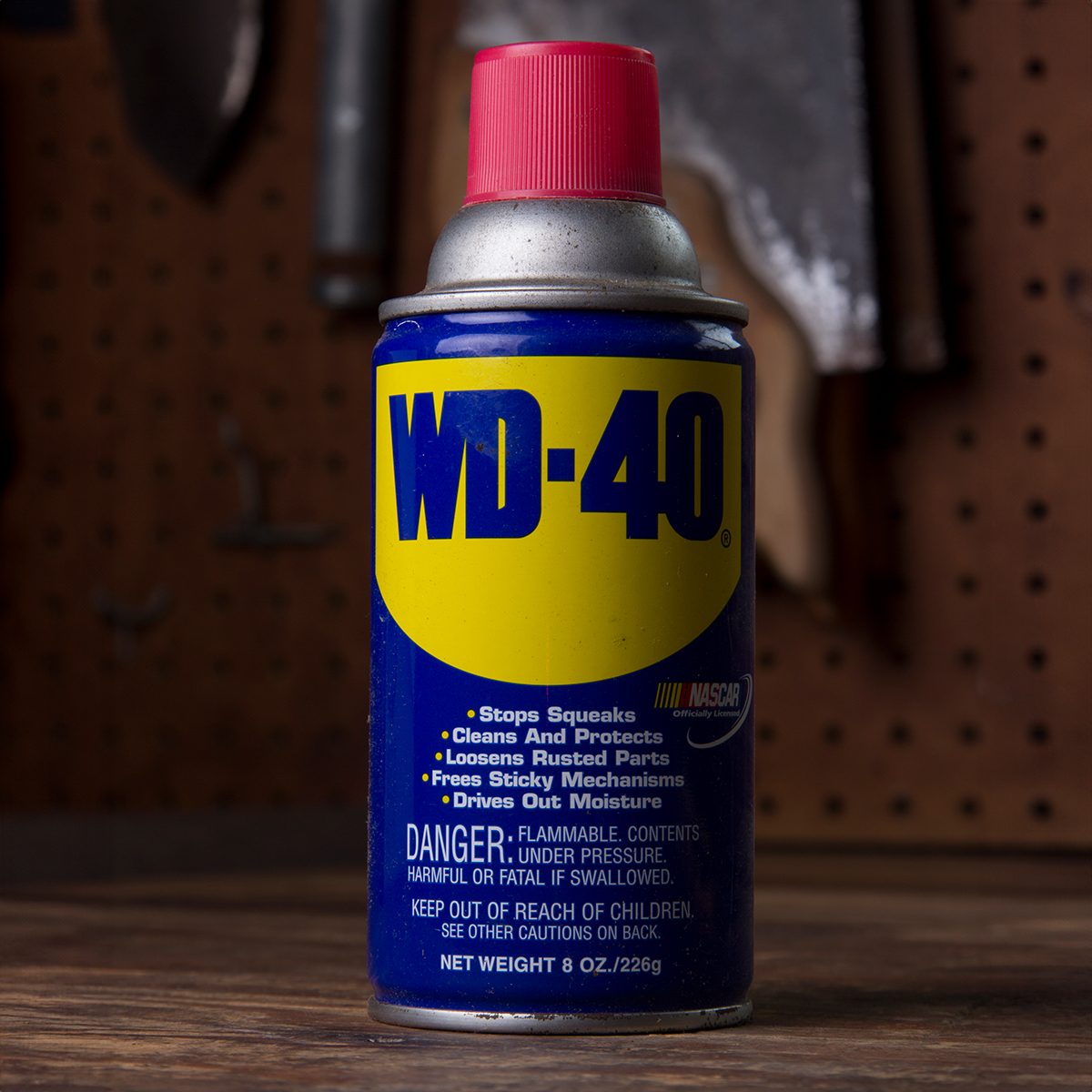 A can of WD-40 lubricant with a blue and yellow label and red cap sits on a workbench, with tools hanging on a pegboard in the background.
