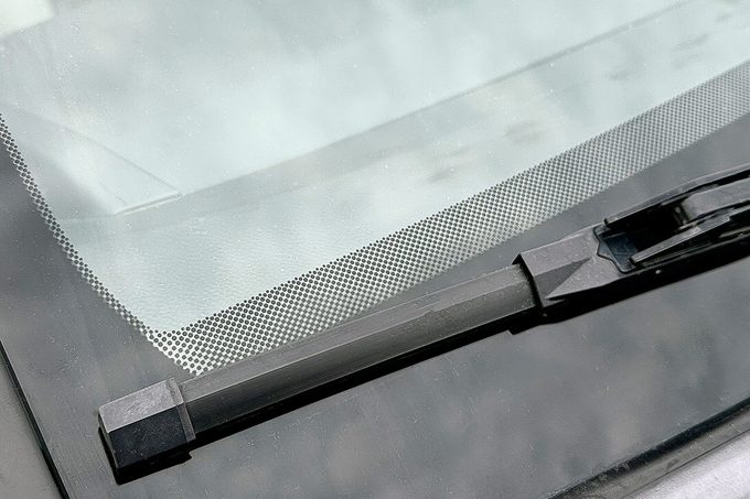 Close-up view of a car windshield with a windshield wiper. The glass is clear, showing a reflection of the sky and clouds above. The wiper blade rests at the base of the windshield.