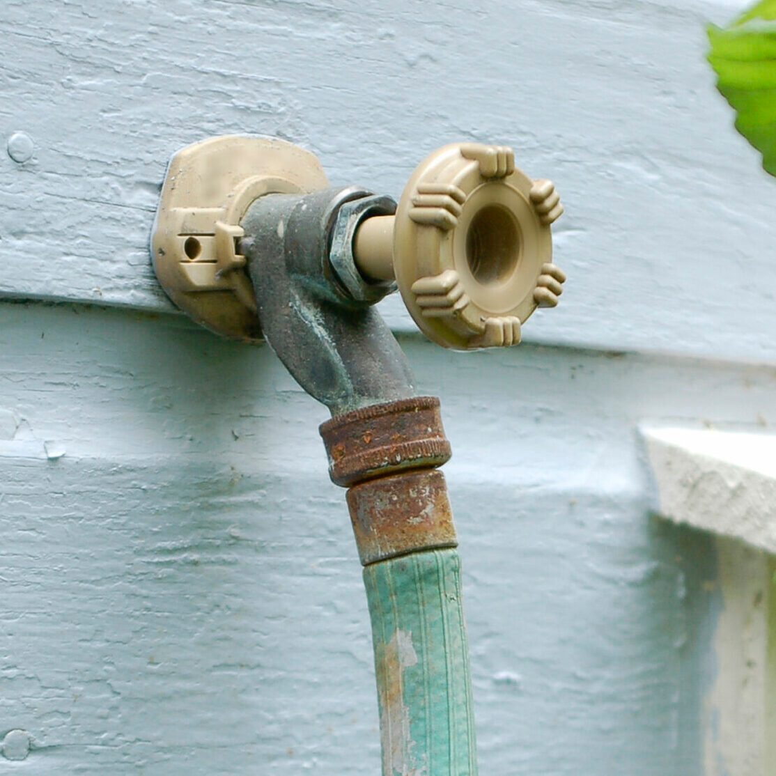 A close-up of an outdoor water spigot with a beige knob, attached to a weathered green garden hose against a light blue wooden wall.