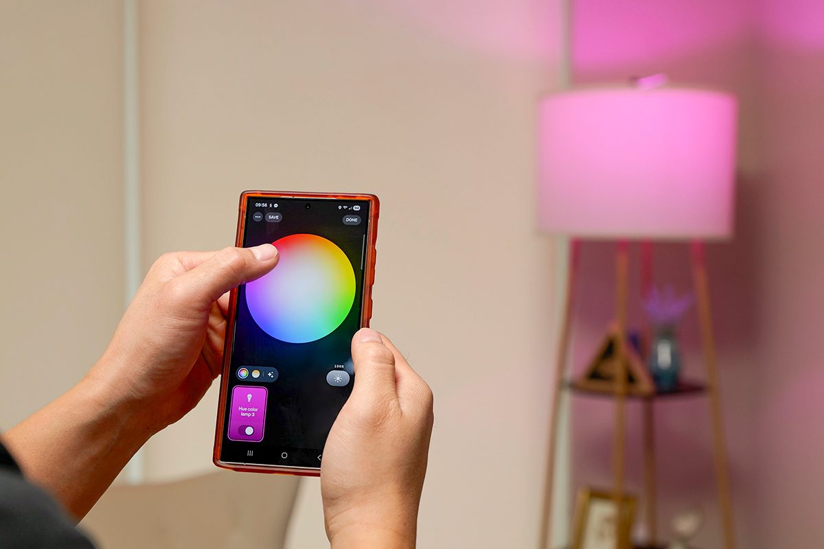 A person uses a smartphone app to adjust the color of a smart bulb, changing a lamp’s light to pink in a modern living room.