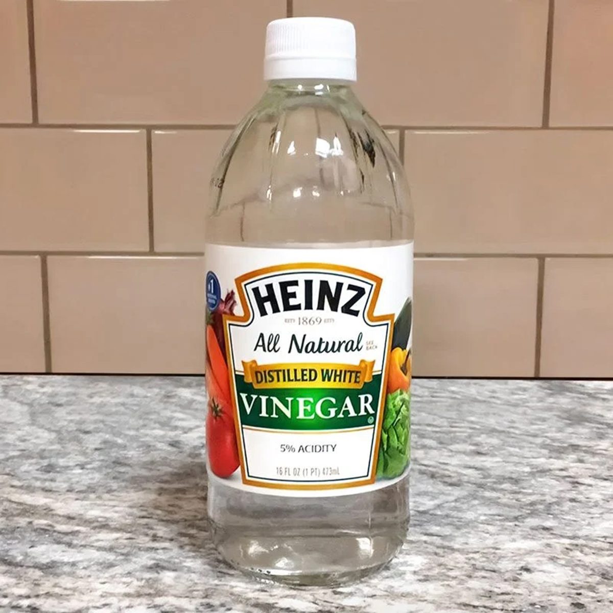 A clear plastic bottle of Heinz distilled white vinegar with a white cap sits on a gray countertop in front of a beige tiled wall. The label features images of tomatoes and vegetables.